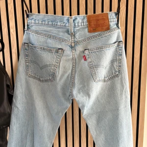 Levi's 501 ljusblå jeans W27 L28 - Klassiska Levi's 501 jeans i ljusblå tvätt med raka ben och femficksmodell. Jeansen har knappgylf, kopparfärgade nitar och den ikoniska läderpatchen bak i midjan. Perfekta för en avslappnad och tidlös stil.