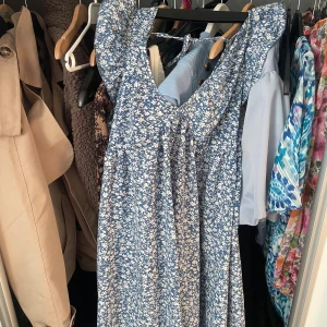 Blå blommig långklänning med volang - Supersöt blå långklänning med vitt blommönster och volangdetaljer vid axlarna. Klänningen har v-ringning och är ärmlös, perfekt för varma dagar. Lätt och flowy modell som ger en romantisk vibe.