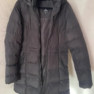 Svart pufferjacka från Threadbare strl 36 - Svart lång pufferjacka från Threadbare med huva och dragkedja framtill. Jackan har två snedställda fickor med dragkedja och quiltad design. Perfekt för kalla dagar och har en stilren look som funkar till det mesta.