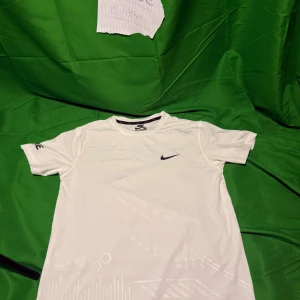 Vit Nike t-shirt  - Snygg vit t-shirt från Nike med diskret mönster och svart swoosh-logga på bröstet. Tillverkad i ett lätt och svalt funktionsmaterial, perfekt för sport eller träning. Klassisk passform med rund hals och korta ärmar. Nike-tryck på ena ärmen.Liknande modell som originalet!