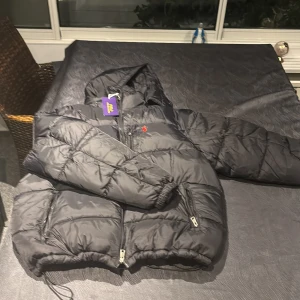 Svart pufferjacka Polo Ralph Lauren M - Svart pufferjacka från Polo Ralph Lauren i storlek XL men passar som M. Jackan har huva, två fickor med dragkedja och klassisk röd broderad logga på bröstet. Tillverkad i polyester och perfekt för kalla dagar. Snygg och stilren design med quiltade partier. Priset kan diskuteras. Först till kvarn