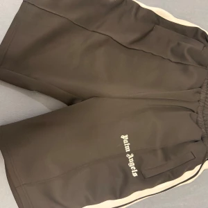 Svarta Palm Angels shorts med vit stripe - Säljer ett par svarta shorts från Palm Angels med vit sidostripes och logga framtill. De har elastisk midja och är gjorda i mjuk polyester, perfekt för en sportig och trendig look. Klassisk loose fit och riktigt snygga detaljer.