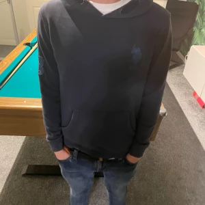 Mörkblå hoodie från USPA - Säljer en mörkblå hoodie från USPA med broderad logga på bröstet. Tröjan har huva, känguruficka och långa ärmar. Skön passform och klassisk stil, perfekt till jeans eller joggers.