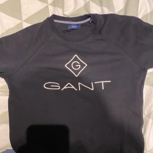 Svart sweatshirt från GANT - Svart sweatshirt från GANT med stort broderat logotyptryck på bröstet. Tröjan har rund halsringning och långa ärmar med ribbade muddar. Perfekt för en chill och stilren look.