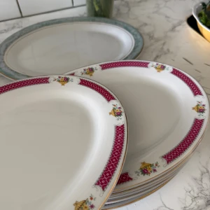 Tallrikar - Bilderna visar en uppsättning ovala porslinstallrikar med vackert mönstrad bård i rött, guld och blommor. Tallrikarna ser ut att vara i gott skick utan synliga nagg eller sprickor. Perfekt för dig som vill duka elegant till fest eller vardag.