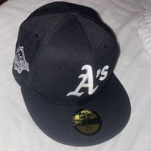 New Era Oakland Athletics keps - Säljer en svart New Era 59FIFTY keps med Oakland A's logga framtill och broderad 1989 World Series patch på sidan. Kepsen är tillverkad i 100% ull och har klassisk rak skärm. MLB-logga baktill och detaljer i vitt. Den är 58.7cm