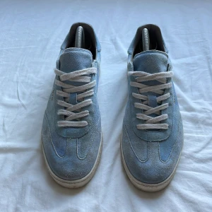 Blå mocka sneakers från AllSaints - Snygga ljusblå sneakers från AllSaints i mjuk mocka med vita snören och vit platt sula. Skorna har en stilren design med diskret guldtext på sidan och klassisk rund tå. Perfekta för dig som gillar en clean och modern look. 