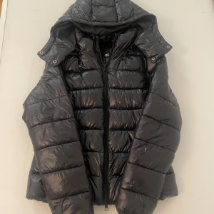 Svart pufferjacka från 157 med huva - Svart pufferjacka från 157 i storlek S. Jackan har en glansig finish, avtagbar huva med tryckknappar och dragkedja framtill. Quiltad design med breda kanaler och lång ärm. Perfekt för kalla dagar och enkel att styla till streetwear.