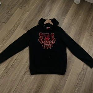 Svart Kenzo hoodie med tigerbrodyr XL - Svart hoodie från Kenzo i storlek XL med ikonisk tigerbrodyr i rött och rosa på bröstet. Tröjan har huva med snörning och en klassisk känguruficka framtill. Perfekt statement-plagg för dig som vill sticka ut.