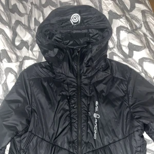 Svart pufferjacka från Sail Racing - Svart pufferjacka från Sail Racing med huva och dragkedja framtill. Jackan har en sportig look med logga på bröstet och är tillverkad i glansig syntet. Perfekt för kalla dagar när du vill ha både stil och värme.