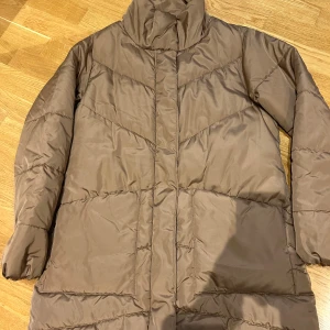 Brun täckjacka från KappAhl - Säljer en brun pufferjacka från KappAhl i storlek S. Jackan har hög krage, långa ärmar och stora fickor framtill. Tillverkad i 100% polyester och har ett stilrent, quiltat mönster. Perfekt för kalla dagar när du vill hålla dig varm och snygg.