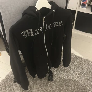 Svart hoodie Maseme med strasstryck - Svart hoodie från Masteme i storlek XS med dragkedja och huva. Framsidan har ett coolt strasstryck med text och baksidan har en stor bild av ett ansikte med spelkort i rött och silver. Perfekt för dig som gillar streetstyle och unika detaljer. Storleken passar S. Väldigt lite använd. 
