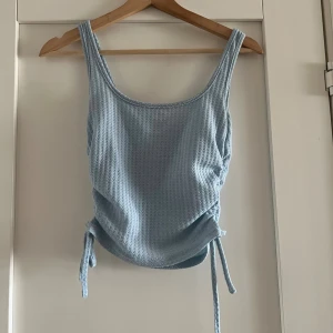 Ljusblå ribbad linnetopp med knyt S - Supersöt ljusblå linnetopp med ribbad struktur och knyt i sidorna. Toppen har breda axelband och en rundad halsringning. Storlek S