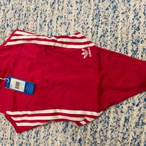 Rosa Adidas bodysuit med vita ränder XS - Snygg rosa bodysuit från Adidas med klassiska vita ränder längs sidorna och Adidas-logga framtill. Modellen är tight och har tryckknappar i grenen. Perfekt för stranden eller poolen, riktigt sportig vibe!