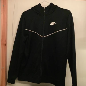 Svart Nike hoodie med dragkedja s - Svart hoodie från Nike i storlek s med vit logga på bröstet och hel dragkedja. Tröjan har huva, långa ärmar och en diskret detalj i form av ett band över bröstet. Perfekt för en sportig och avslappnad stil.
