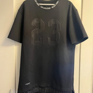 Svart t-shirt Cayler & Sons Black Label XL - Svart t-shirt från Cayler & Sons Black Label i storlek XL. Tryck med siffran 23 på ryggen och texten LEGEND nedtill. Rund hals med logotypband och korta ärmar. T-shirten har en clean streetwear-look och är tillverkad i mjukt material.