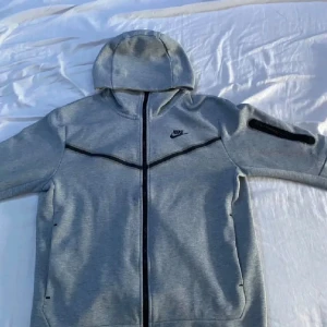 Grå Nike hoodiejacka med dragkedja - Snygg grå hoodiejacka från Nike med hel dragkedja framtill och svarta detaljer. Jackan har huva, två sidofickor och en extra ficka med dragkedja på ärmen. Perfekt för en sportig och avslappnad stil.