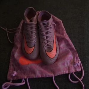Nike Mercurial lila fotbollsskor elite - Säljer ett par Nike Mercurial fotbollsskor i lila med orange detaljer och svart swoosh. Skorna har snörning, platt sula med fasta dobbar och är tillverkade i syntetmaterial. Perfekta för dig som vill sticka ut på planen. Levereras med matchande lila Nike-påse.