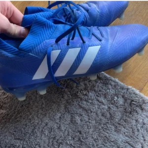 Blå adidas fotbollsskor med snörning - Säljer ett par blå fotbollsskor från adidas med de klassiska vita ränderna på sidan. Skorna har snörning och är gjorda i syntetmaterial med mönstrad yta för extra grepp. Perfekta för dig som vill ha stil på planen.