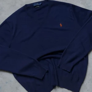 Marinblå tröja från Polo Ralph Lauren - Snygg marinblå långärmad tröja från Polo Ralph Lauren i slim fit. Tillverkad i mjuk pima bomull och pryds av den klassiska orangea polospelaren broderad på bröstet. V-ringad hals och stilren design som passar perfekt till jeans eller chinos.