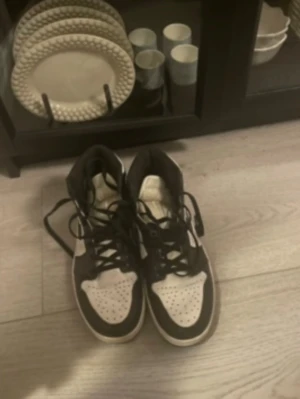Svarta och vita Nike Jordan 1 sneakers - Säljer ett par klassiska Nike Jordan 1 sneakers i svart och vitt. Skorna har snörning, perforerad tå och högt skaft. Perfekta för dig som gillar streetwear och vill ha en ikonisk sneaker med snygg kontrast mellan färgerna.