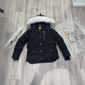 Svart dunjacka Moose Knuckles med päls - Svart dunjacka från Moose Knuckles med fluffig vit päls på huvan, silverfärgade detaljer och dragkedjor. Jackan har flera fickor med knappar, ribbade muddar och ett snyggt broderat motiv på insidan. Perfekt för kalla dagar och riktigt stilren look.