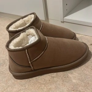 Bruna fodrade boots med platt sula - Mysiga bruna boots med platt sula och rund tå. Skorna har ett varmt, vitt foder på insidan och är perfekta för kyliga dagar. Utsidan är i mockaliknande material och de har dekorativa sömmar runt skaftet.