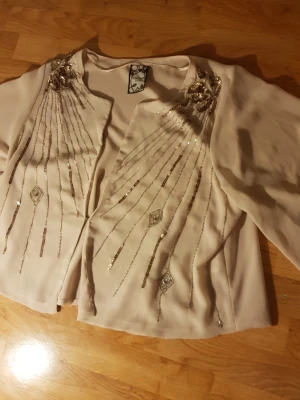 Beige kofta med paljetter Vintage Stories - Snygg beige kofta från Vintage Stories i storlek 40/42. Koftan har långa ärmar och är dekorerad med glittriga paljetter och broderade detaljer i silver som strålar ut från axlarna. Perfekt för dig som vill sticka ut med en unik och glamorös look.