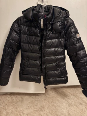 Svart Moncler pufferjacka med huva - Säljer en svart moncler jacka då jag inte använder den längre. Jag använde den endast under en vinter så den är i väldigt bra kvalité. Det enda felet är att tygbiten med cartoon bilden har gått av lite men det går bara att sy på igen enkelt! Den är storlek 00 men passar även xs-s