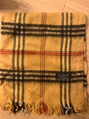 Rutig beige väst från Burberrys - Säljer en klassisk rutig väst från Burberrys i beige med svarta, vita och röda ränder. Västen är tillverkad i 100% lammull och har fransar längst ner. Perfekt för dig som gillar tidlös design och vill ha något som sticker ut.