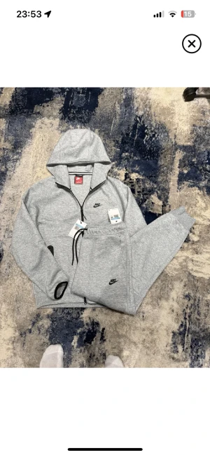 Nike tech tracksuite helt ny grå - Säljer ett par grå mjukisbyxor från Nike med svart logga på benet. Byxorna har snörning i midjan, ribbade muddar vid benslut och en svart dragkedja längs ena benet. Materialet är mjukt och skönt, perfekt för chill eller träning.