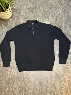 Mörkblå stickad pikétröja från SMOG - Mörkblå stickad pikétröja från SMOG med klassisk krage och half zip-dragkedja framtill. Tröjan har långärmad passform och ribbad struktur som ger en snygg och stilren look. Perfekt för dig som gillar en clean och enkel stil.