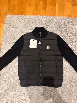 Svart cardigan från Moncler - Säljer en svart pufferjacka från Moncler med stickade ärmar och hög krage. Jackan har quiltad front, dragkedja hela vägen och två fickor med knappstängning. Klassisk Moncler-logga på bröstet och medföljande dustbag. Perfekt för höst och vinter!