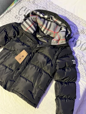 Svart pufferjacka från Burberry - Svart pufferjacka från Burberry med avtagbara ärmar och huva fodrad med klassiskt rutigt mönster i grått, vitt och rött. Jackan har dragkedja, knappar och två fickor med zip framtill. Perfekt för dig som vill ha en snygg och mångsidig jacka.