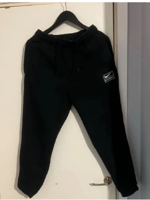 Svarta Stussy x Nike mjukisbyxor - Svarta mjukisbyxor med broderad Stussy x Nike-logga på vänster ben i storlek M. Byxorna har elastisk midja med snörning och är tillverkade i mjukt bomullsmaterial. Passformen är loose och benen är avsmalnande mot ankeln. Perfekta för chill eller streetwear.  