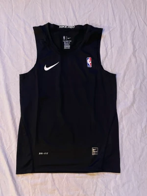 Nike Pro NBA Dri-Fit linne - Snyggt svart linne från Nike Pro med NBA-logga och Dri-Fit-teknologi. Perfekt för träning och sport, med en stilren design och bekväm passform. Materialet är syntet och håller dig torr under intensiva aktiviteter. Nike-logga på bröstet och etikett nedtill.