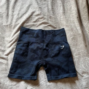 Svarta camo shorts från Gymshark - Säljer ett par svarta högmidjade shorts från Gymshark med mörk camo-mönster. De är tighta och stretchiga, perfekta för träning. Materialet är mjukt och följsamt syntet, med diskret logga framtill. Sitter snyggt och bekvämt på kroppen.