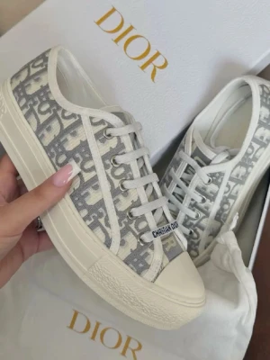 Gråvita Dior sneakers med monogram - Säljer ett par snygga Dior sneakers med grått och vitt monogrammönster. Skorna har rund tå, platt sula och klassisk snörning. Materialet är bomullstyg med vita detaljer och logotypen 'CHRISTIAN DIOR' på plösen. Perfekt för dig som gillar lyxiga streetwear-vibes.