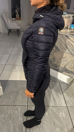 Svart Parajumpers pufferjacka med huva - Säljer en svart pufferjacka från Parajumpers med huva och snygga detaljer. Jackan har dragkedja framtill, två sidofickor med dragkedja och ett broderat märke på ärmen. Passformen är slim och jackan är perfekt för kyliga dagar.