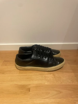  Saint Laurent - Säljer ett par svarta sneakers från Saint Laurent med beige platt sula och snörning. Skorna är i skinn och har en klassisk rund tå. Märket syns på både hälen och undersidan av sulan. Perfekta för dig som gillar stilrena och lyxiga sneakers.