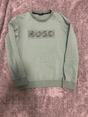 Grön sweatshirt från BOSS med tryck - Snygg grön sweatshirt från BOSS med svart logotyptryck på bröstet. Tröjan har rund hals, ribbade muddar vid ärmslut och nederkant samt långärmad passform. Perfekt för dig som gillar stilrena och bekväma plagg.