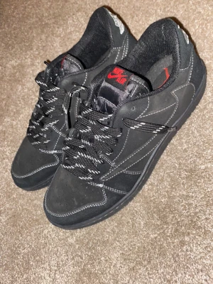 Nike Air Jordan 1 Low Travis Scott svart - Nike Air Jordan 1 Low Travis Scott i svart mocka med vita kontrastsömmar och broderade detaljer på hälen. Skorna har svarta och vita snören, röd Nike Air-logga på plösen och unika Travis Scott-inslag som bee embroidery och text på innersulan.