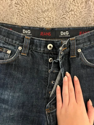 D&G jeans i mörkblå denim wide fit - Säljer ett par mörkblå jeans från D&G. Modellen har breda ben och normal passform, med knappgylf och läderpatch bak i midjan. 