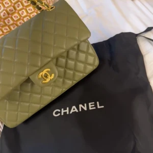 Handväska  - Säljer en khaki färgad handväska med klassiskt quiltat mönster och guldfärgad kedja samt logodetalj framtill. Väskan har en elegant och tidlös design, perfekt för att lyfta din outfit! Dustbagen är tyvärr borta men ska försöka leta fram den💞