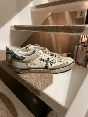 Premiata vita sneakers med blå detaljer - Säljer ett par vita Premiata sneakers med blå detaljer och logga på sidan. Skorna har snörning, rund tå och platt sula. Materialet är skinn och de har en cool, lite vintage look med mönstrad häl. Perfekt för dig som gillar stilrena och unika sneakers.
