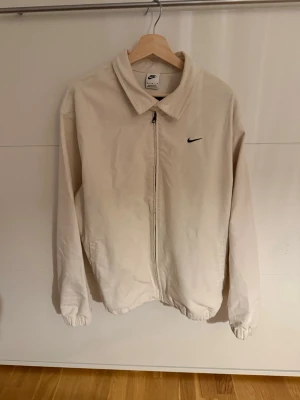 Beige Overshirt från Nike - Snygg beige vindjacka från Nike med klassisk krage och svart logga på bröstet. Jackan har dragkedja framtill, elastiska muddar vid ärmslut och nederkant samt två sidofickor. Perfekt för dig som gillar stilren och enkel design.