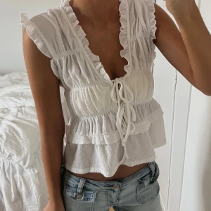 Vit blus med volanger och snörning - Säljer en söt vit blus med volanger och djup v-ringning. Toppen har snörning framtill och är croppad med flera lager volanger. Materialet är lätt och luftigt, perfekt för varma dagar. Ärmlös design med romantiska detaljer som ger en fräsch och trendig look.