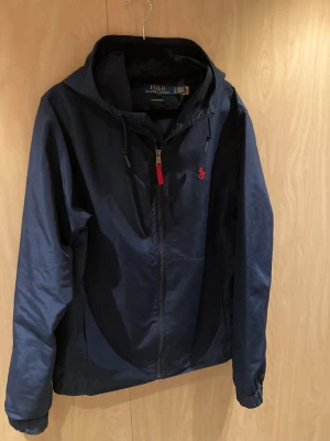 Ralph Lauren vindjacka - Snygg marinblå vindjacka från Polo Ralph Lauren med huva och röd logga på bröstet. Jackan har dragkedja med rött band, två sidofickor och elastiska muddar. Perfekt för dig som gillar stilren och sportig look.