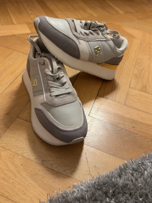 Grå och beige sneakers med gul detalj - Snygga sneakers från Tommy Hilfiger i grått och beige med gul detalj på sidan och guldfärgad logga. Skorna har snörning, rund tå och platt sula. Materialmix av skinn och mocka med textilpaneler för extra stilpoäng.