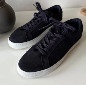 Marinblå sneakers från Les Deux - Snygga marinblåa sneakers från Les Deux med ovandel i mocka och vita platta sulor. Skorna har snörning och broderade detaljer i blått, vitt och rött på hälen. Perfekta för dig som gillar stilrena och klassiska sneakers med en twist.